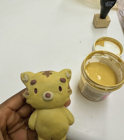 A hand-sculpted clay tiger figurine held beside a jar of yellow glaze.